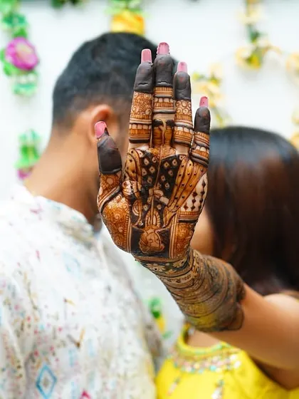 A creative shot of the bride's mehendi, with the groom in the background. The focus is on the detailed figures on her palm.