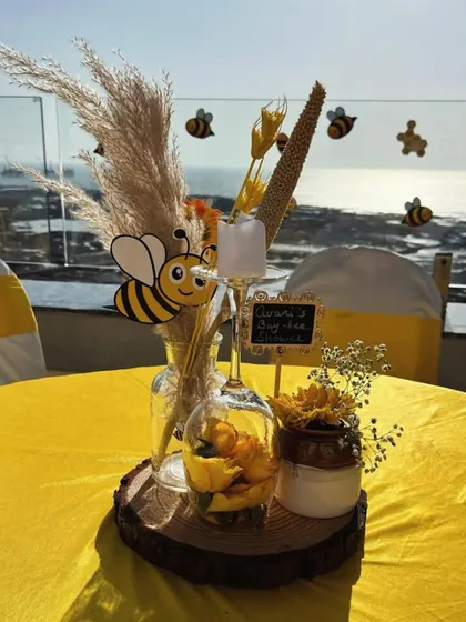 A close-up of a tabletop centerpiece for our bee-themed baby shower. This arrangement sits on a rustic wood slice and includes pampas grass, a cute bee cutout, yellow flowers, and a small chalkboard sign, all set against a beautiful seaside view.