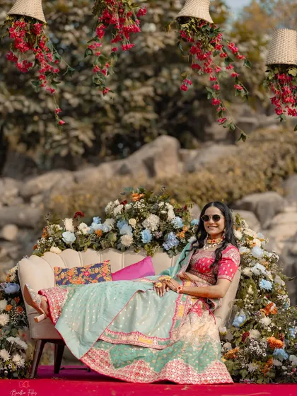 The bride looking radiant and relaxed on a sofa adorned with flowers during her outdoor Mehendi ceremony.