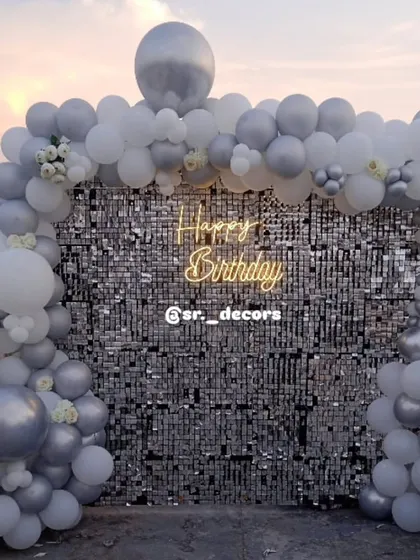 We take the party anywhere, even your rooftop. This setup features a brilliant silver shimmer wall framed by a white and silver balloon arch, creating a chic and elegant photo corner for an outdoor celebration.