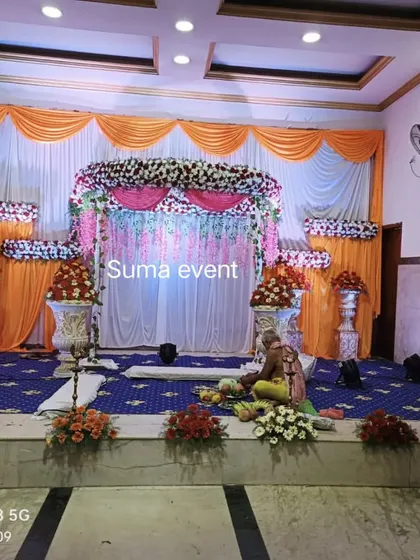 A traditional stage decoration for a naming ceremony in a function hall. The setup includes a central floral arrangement, decorative urns with flowers, and bright orange and white drapes.