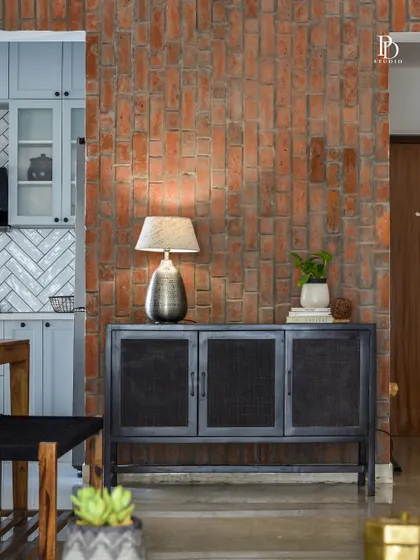 An exposed brick wall adds raw, earthy texture to this dining area, creating a beautiful contrast with the sleek, modern kitchen beyond. The black metal and mesh sideboard provides stylish storage while maintaining a light, industrial feel.