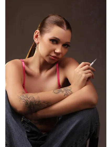 A bold portrait of a model with tattoos, wearing a pink lace bralette and smoking. The direct gaze and confident posture create a rebellious and empowered look.