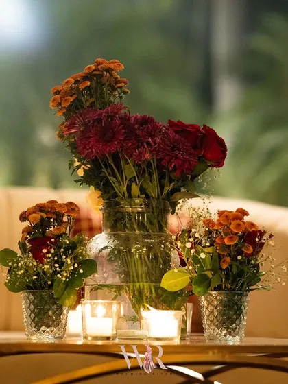 A close-up of a rustic yet elegant centerpiece. A simple glass jar holds a bouquet of red and orange flowers, perfect for the warm, earthy theme of the wedding.