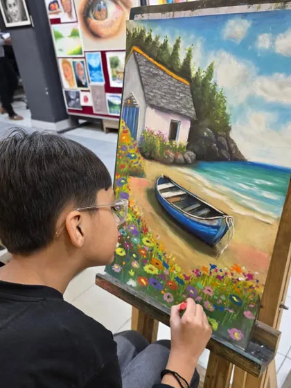 Another angle of the young artist at his easel. He is using a reference photo to guide his drawing of the boat and the shoreline.