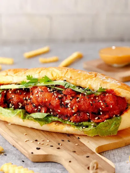 A loaded Korean-style chicken sandwich on a wooden board, surrounded by scattered fries.