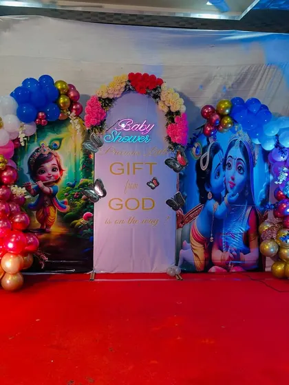 An elegant and premium baby shower decoration with a Krishna theme. The stage is beautifully lit with colored lights, enhancing the vibrant balloon arches and backdrops.