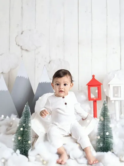 A cozy 'Winter Wonderland' themed session. This little one is dressed in a simple white outfit, sitting amongst fake snow, miniature pine trees, and mountain cutouts.