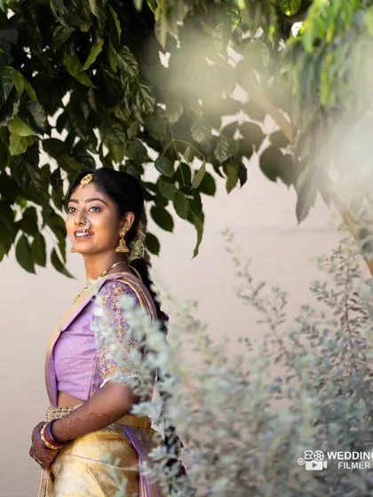 A beautiful outdoor shot. The natural light highlights the soft, glowing makeup and the delicate details of her lavender silk saree, perfect for a daytime traditional event.