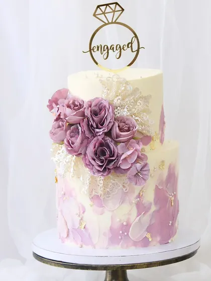 A sophisticated two-tiered engagement cake with a beautiful lavender and white watercolor effect. The design is elevated with intricate lace details, gold leaf accents, and a cluster of deep purple sugar roses.
