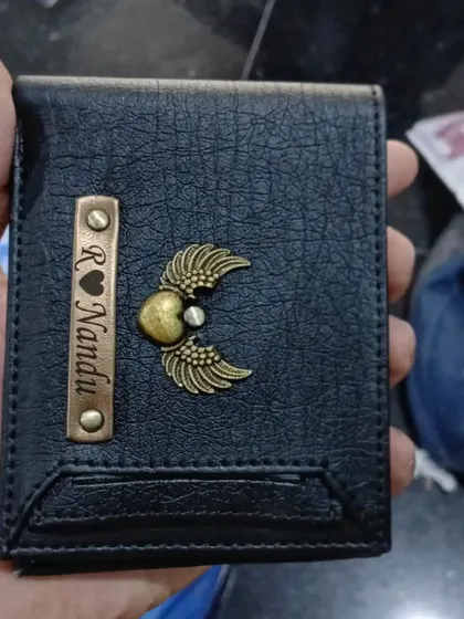 A personalized black wallet with a heart and wings charm, held to show the texture and craftsmanship.