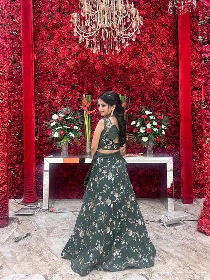 A happy client rocking a dark green floral print lehenga set against a stunning red flower wall. This shows how our outfits are perfect for any grand celebration.