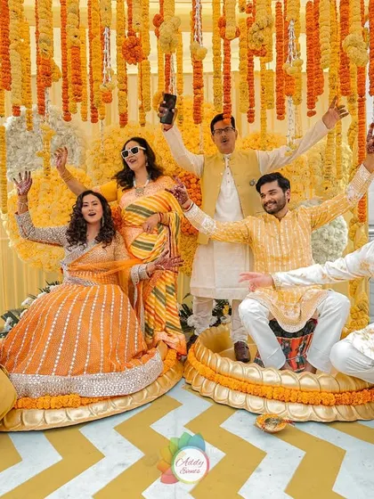 A fun group photo from the Haldi ceremony, with the couple and their friends posing joyfully against the vibrant marigold backdrop.