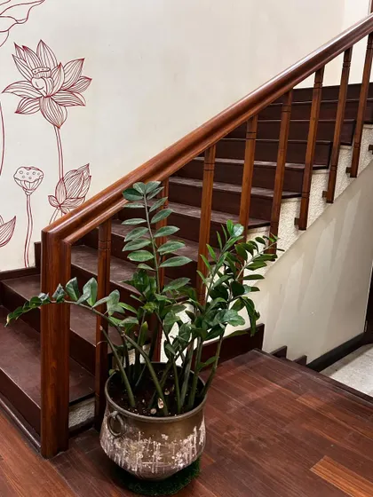 A hardy ZZ plant in a rustic, antique-finish metal pot sits gracefully at the bottom of a wooden staircase. The lotus-themed wall art behind it complements the natural, tranquil aesthetic.