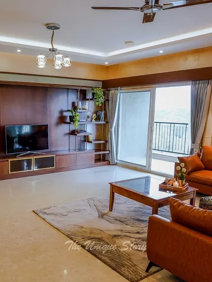 The full view of the living room highlights the warm and inviting atmosphere created by the tan leather sofas and extensive wooden TV unit. The large sliding doors open to a balcony, flooding the room with natural light.
