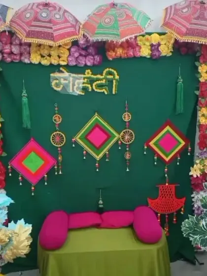 A Mehendi backdrop with a dark green curtain, decorated with colorful umbrellas, hanging props, and floral arrangements on the sides.