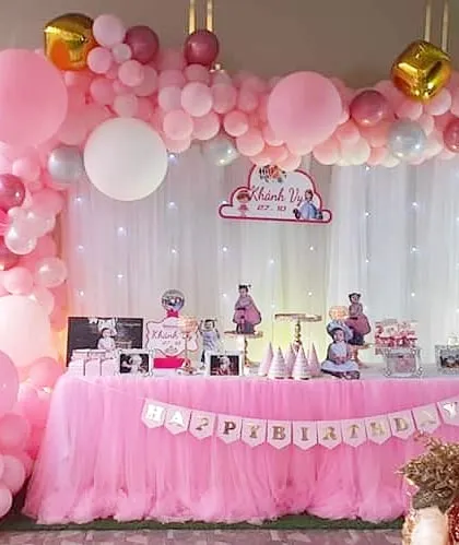 A sweet and personal pink-themed decoration. This setup features a beautiful balloon garland over a fairy-lit curtain and includes framed photos of the birthday girl on the main table.