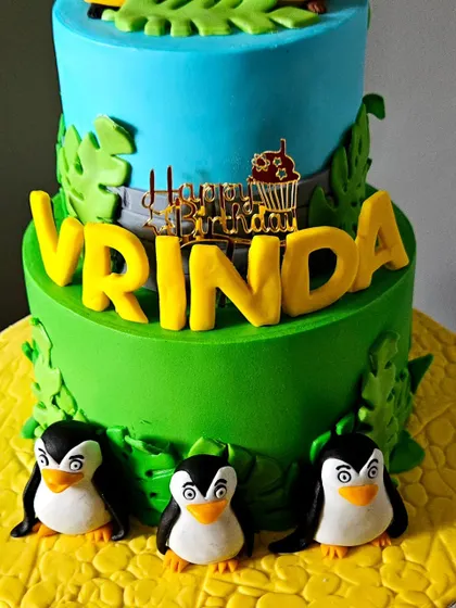A close-up of the base of the Madagascar cake, showing the adorable, hand-sculpted penguin figurines and the bold, 3D name of the birthday girl.