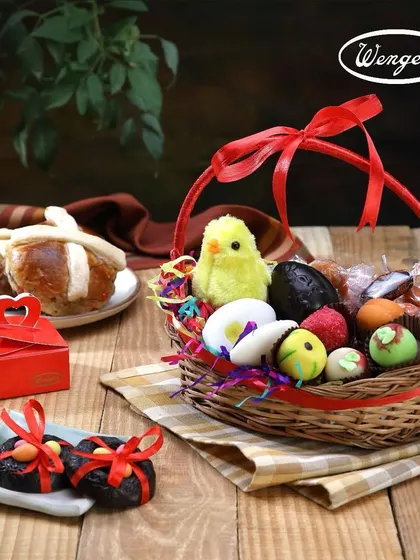 An adorable Easter basket filled with goodies. It includes marzipan eggs, chocolates, a small toy chick, and our famous Hot Cross Buns in the background.