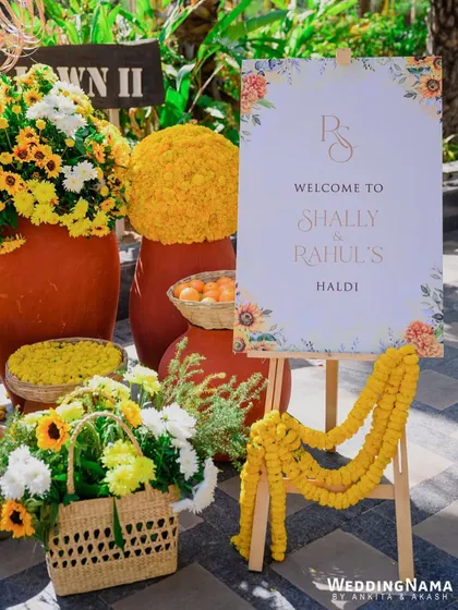 A welcoming entrance for a haldi, with an easel sign surrounded by earthy pots, marigold garlands, and baskets of fresh flowers.