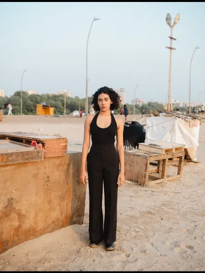 A full-length portrait with a raw, almost documentary feel. The industrial beach background contrasts with the model's simple, chic outfit.
