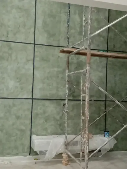 A green-toned textured wall with black grid lines, shown during the application process with scaffolding, achieving a unique tiled appearance.