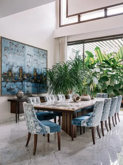 A dining room with a view. The double-height windows not only flood this space with natural light but also make the lush garden a living backdrop for every meal. The blue chairs echo the colors in the artwork, creating a cohesive design.