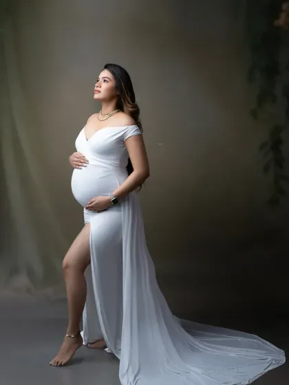 A classic and elegant maternity pose. The simple white gown with a long train accentuates the beauty of the baby bump against a soft, neutral background.