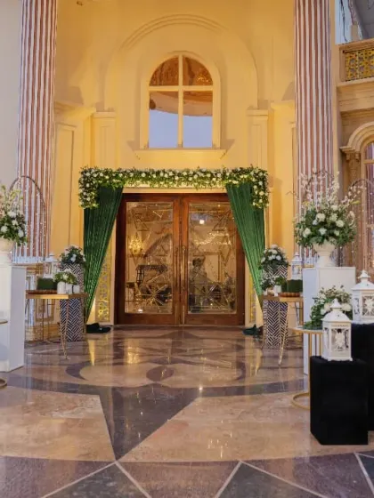 A grand welcome to a palatial venue, with the entrance doors framed by lush green and white floral arrangements. The combination of classic architecture and fresh flowers creates a timeless and elegant first impression.