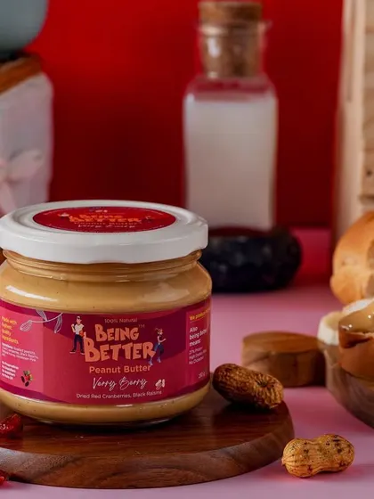 A jar of Being Better's peanut butter, styled with fresh bread and nuts against a bright red background. The composition is energetic and appetizing, highlighting the product as a delicious and healthy spread.