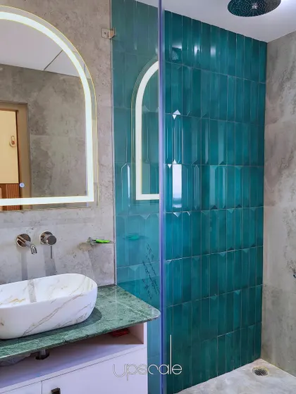 This bathroom design features a striking teal tile in the shower area as a focal point. The rich color is balanced by a green marble countertop and a modern, arched backlit mirror.