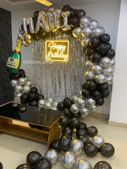 A chic birthday party with a black and silver theme. The circular balloon arch is decorated with a champagne bottle foil balloon and a 'Happy Birthday' neon sign.