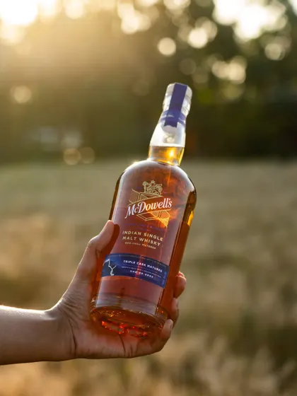 A bottle of McDowell's single malt whisky held up against a golden-hour sunset in a field. This outdoor shot creates a feeling of nature and craftsmanship.