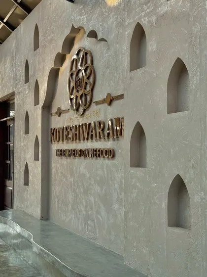 The entrance wall of the Koteshwaram restaurant, featuring traditional arched niches and a metallic logo. The textured concrete finish provides a modern backdrop for these classic design elements.