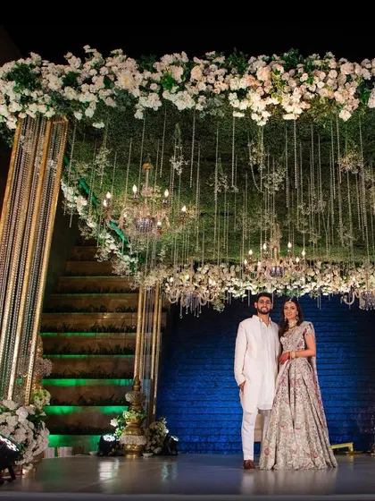 A stunning stage setup for a wedding reception, featuring a canopy of white flowers and hanging crystal chandeliers. This design creates a feeling of grandeur and romance, perfect for the happy couple.
