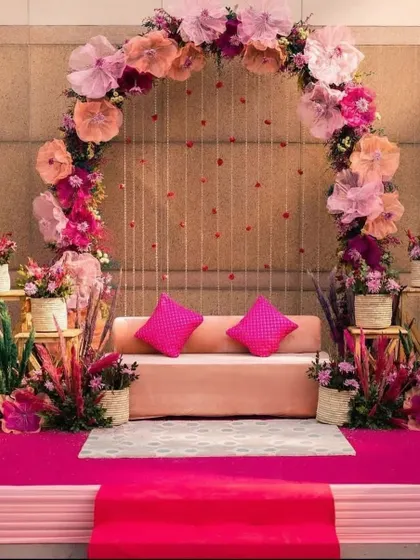 A pretty pink stage setup for a Mehendi or Roka ceremony. The decor features a large arch of paper flowers and a simple, elegant seating arrangement.