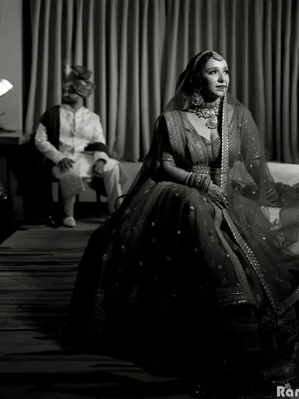 A dramatic black and white portrait with the bride in the foreground and the groom seated in the background, creating an artistic and moody composition.