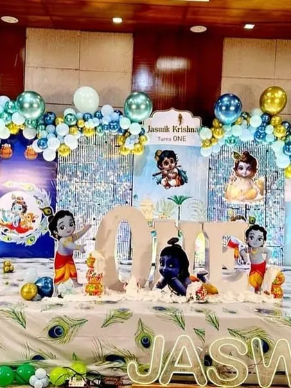 A beautiful Krishna-themed first birthday party. The stage is decorated with peacock feather motifs, multiple Krishna cutouts, and a blue and gold balloon arch.