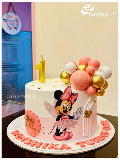 A sweet Minnie Mouse cake for a first birthday. This design features a fairy Minnie illustration, a plaque with the baby's name, and a cascade of pink and gold balloons.