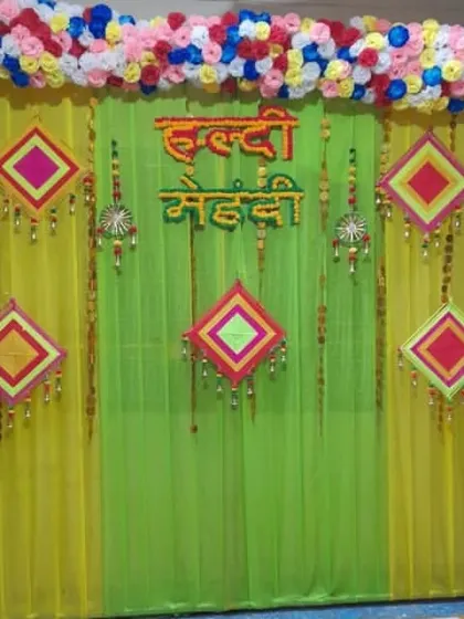A simple and colorful backdrop for a combined Haldi and Mehendi function at home. The decor uses light green and yellow drapes with hanging props.