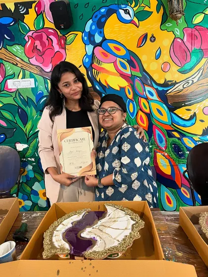 A participant receives her certificate in front of our vibrant peacock mural, her custom resin art piece beside her.