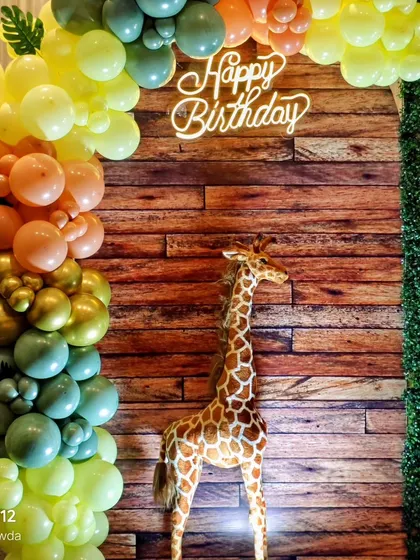 A close-up of a jungle-themed balloon arch with a tall giraffe prop and a neon 'Happy Birthday' sign against a wooden backdrop. This combination of rustic and modern elements creates a unique look.