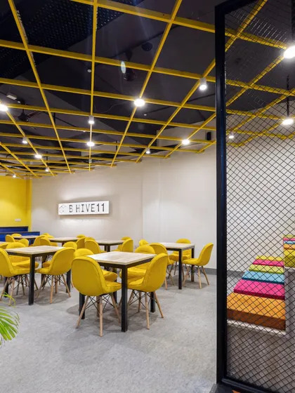 The B-Hive 11 cafeteria features bright yellow chairs and a matching yellow grid on the ceiling, creating a cohesive and energetic look. The space is designed for community, with flexible seating for dining and informal gatherings.