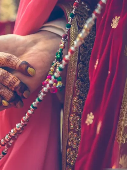 A close-up detail shot of hands during a wedding ritual. The colorful beads and intricate henna patterns highlight the rich textures and traditions of an Indian wedding.