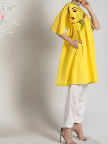 A full-length view of a bright yellow hand-painted kurti paired with white pants. The pop-art style face on the shoulder adds a modern and fashionable twist to the traditional silhouette.