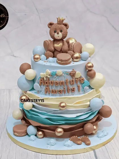 A sweet two-tier "Adventure Awaits" cake to welcome a new baby. This 3kg choco-chip cake features a crowned teddy bear topper amidst blue, brown, and gold decorative balls and macarons.