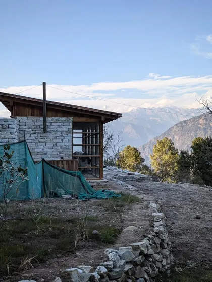A quiet moment at our Uttarakhand site, with the snow-capped Himalayan range in the distance. The building's form, with its simple stone chimney and sloping roof, is a direct response to the climate and the traditional architecture of the region.