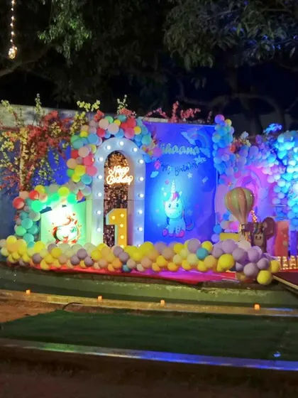 A magical unicorn-themed first birthday party held outdoors at night. The stage is brightly lit and features a pastel rainbow balloon arrangement, a unicorn figure, and a large illuminated number '1'.