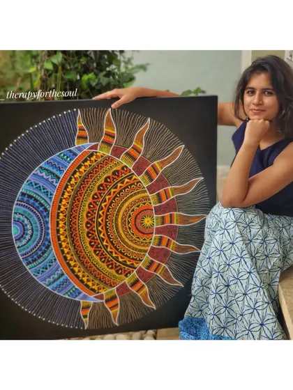 Me sitting with a large, custom sun and moon mandala painted on a black canvas. This piece shows the contrast between warm and cool colors and the intricate details that go into my work.