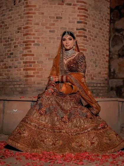 A stunning full-length portrait of the bride seated against a rustic brick wall. The rich brown and gold lehenga is spread out, with scattered rose petals adding a touch of romance to the scene.